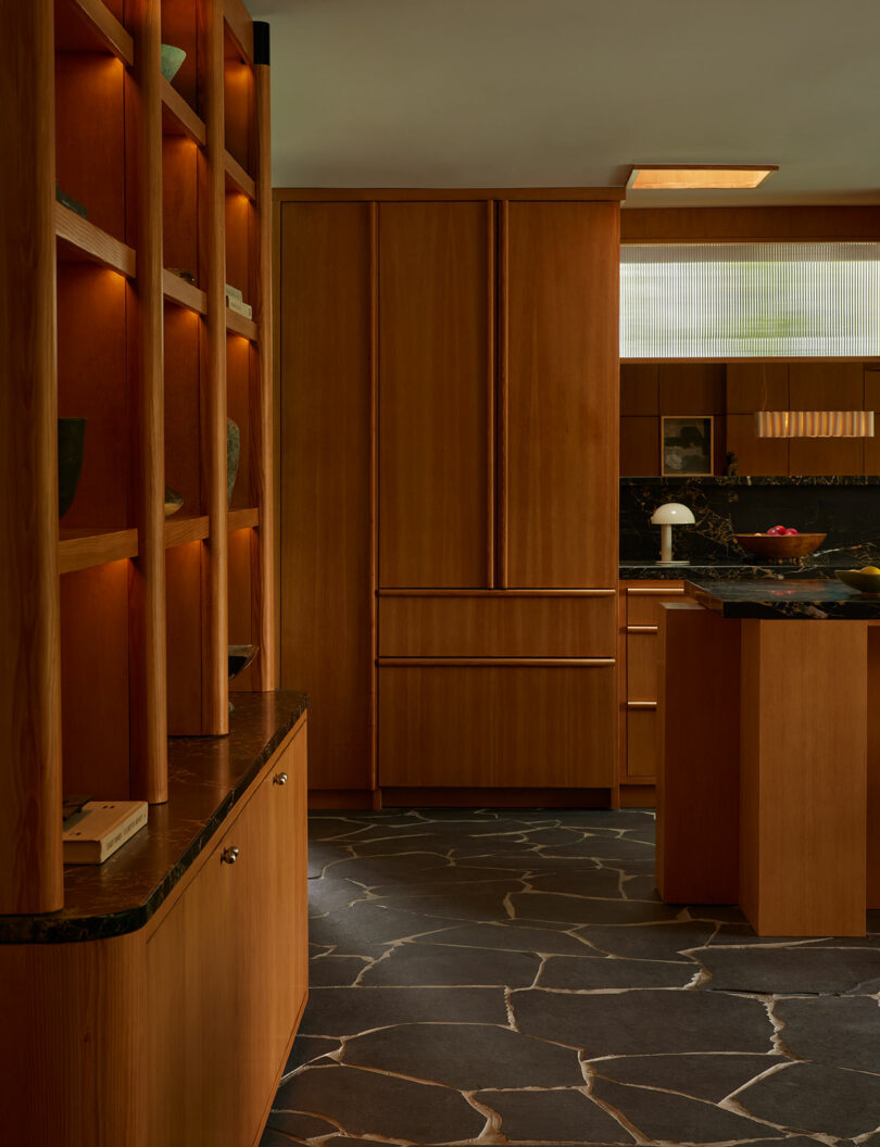 Contemporary kitchen with wood cabinetry, black stone countertops, open shelving, and a dark stone floor with irregular patterns. Warm ambient lighting highlights the cabinets.