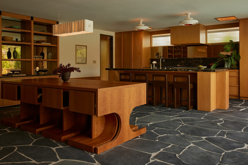 Modern kitchen and dining area with wood cabinetry, a large geometric wood dining table, black stone flooring, and pendant lighting.