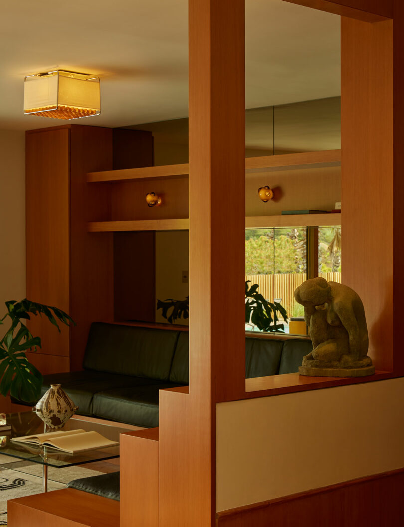 A modern living room with wood paneling, a black sofa, a coffee table with books, plants, a window with outdoor view, and a small sculpture of a seated figure on a shelf.