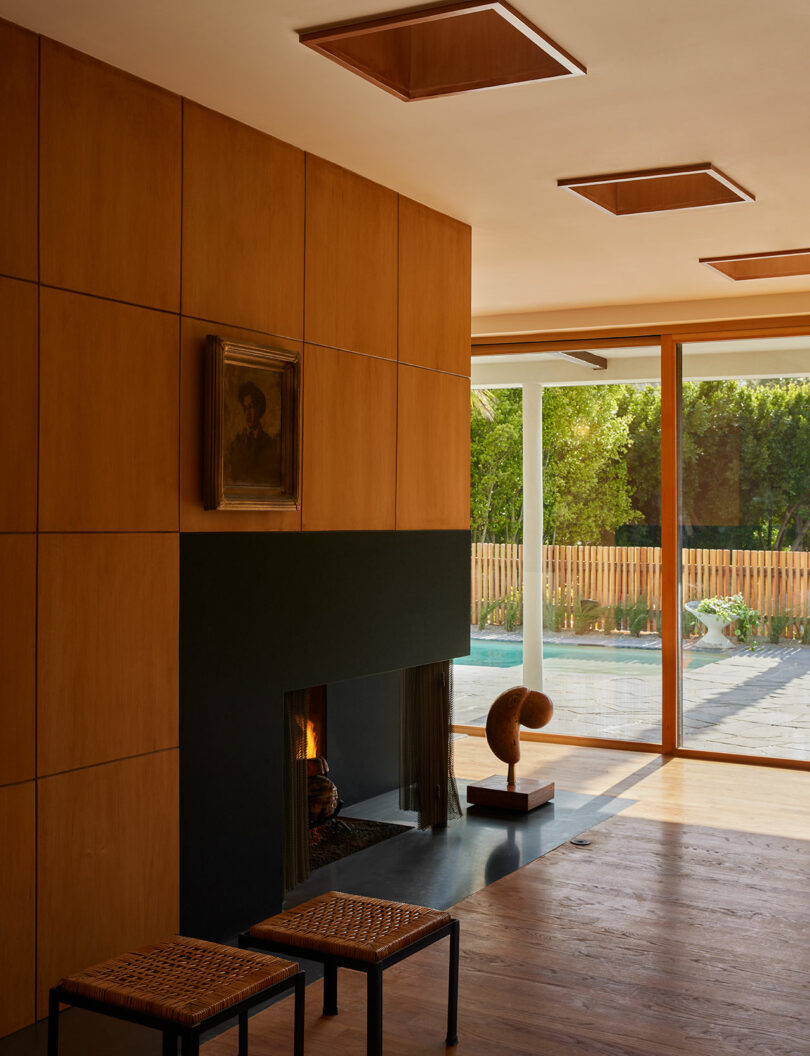 A modern living room with wood-paneled walls, a black fireplace, stools, abstract sculpture, large windows, and a view of a garden outside.