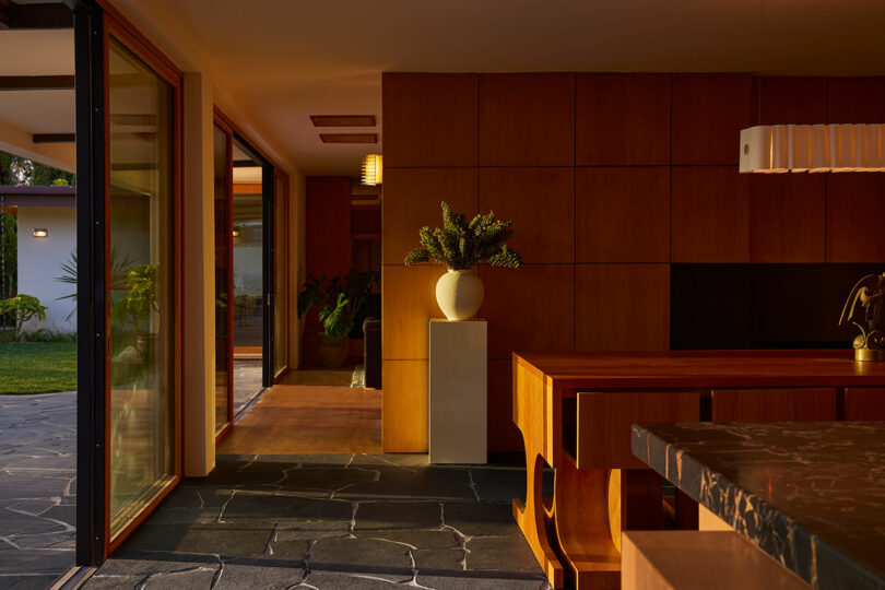 Modern interior with wood-paneled walls, a marble countertop, large sliding glass doors, and a potted plant on a pedestal bathed in warm sunlight.