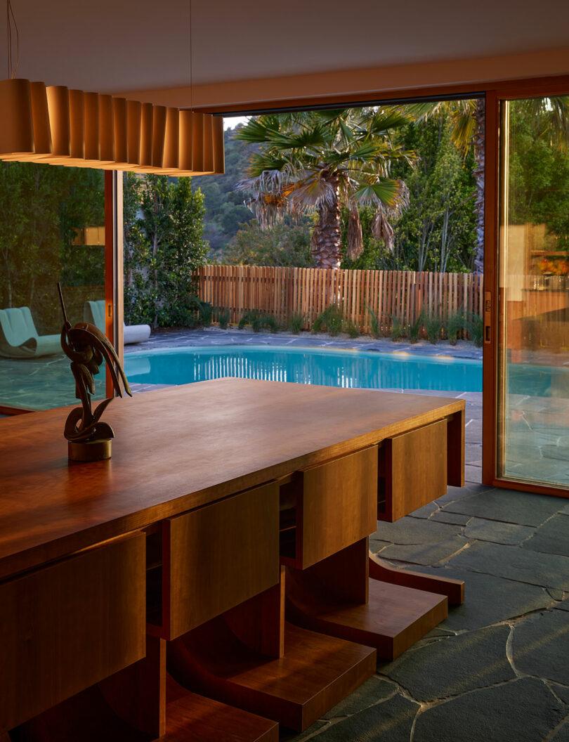 A wooden dining table sits next to an open sliding glass door, overlooking a backyard pool and palm trees, with stone flooring visible indoors.