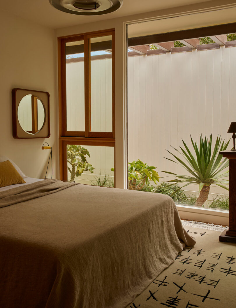 A minimalist bedroom with neutral bedding, a wooden-framed window, a small bedside lamp, and a view of a desert-style garden with succulents through a large glass door.