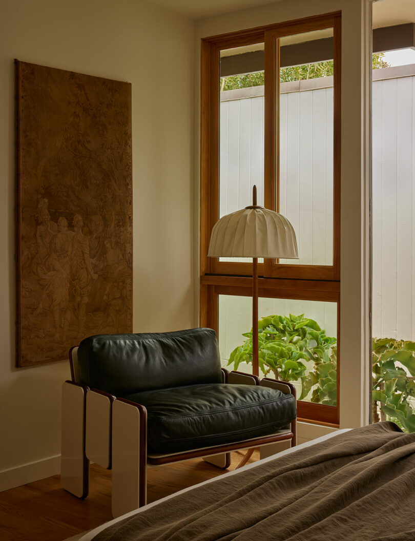 A modern room corner with a black leather armchair, a floor lamp, a wooden-framed window, a large wall art piece, and greenery visible outside.