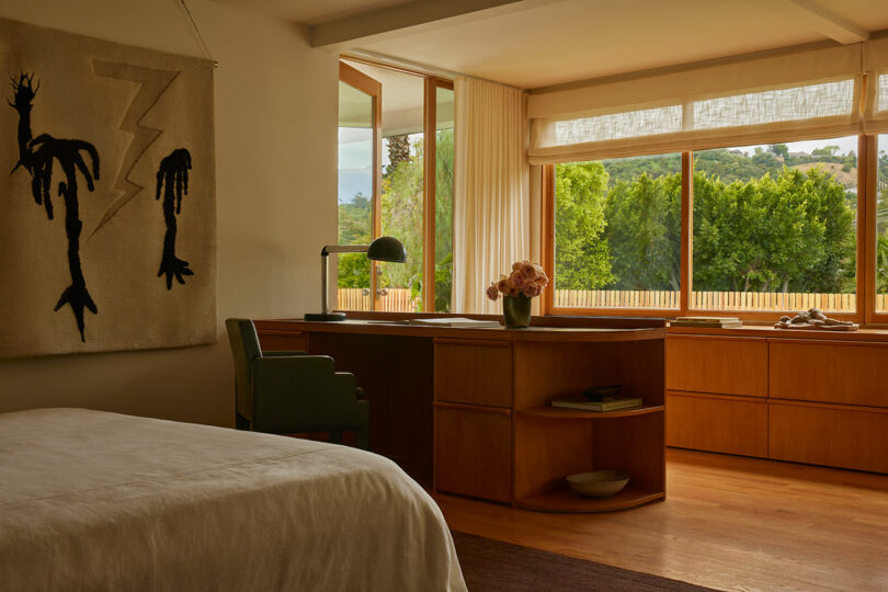 Sunlit bedroom with a bed, wooden desk, chair, and shelves; large windows show green trees and hills outside; abstract wall hanging on the left.