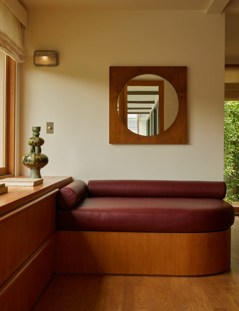 A maroon leather bench with wooden base sits under a round mirror on the wall; a sculptural vase is placed on the adjacent wooden ledge near a window.