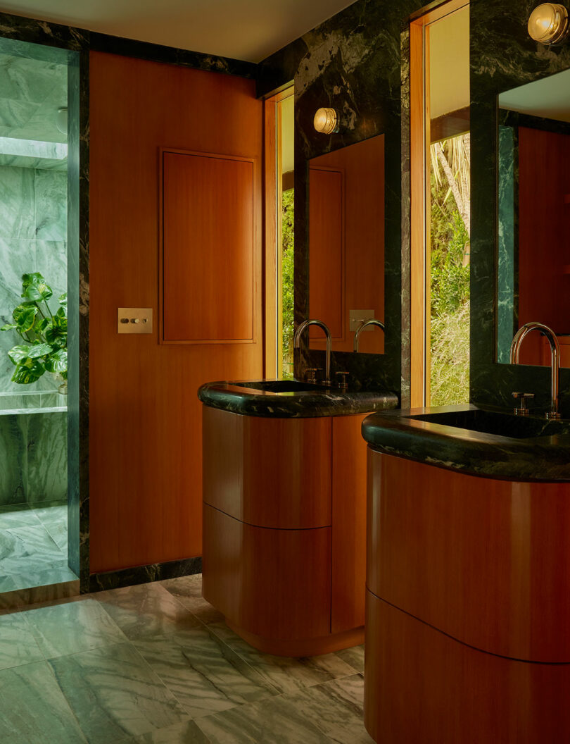 Contemporary bathroom with two wood vanities, black marble countertops, wall mirrors, and large windows overlooking greenery; shower area with marble walls visible on the left.