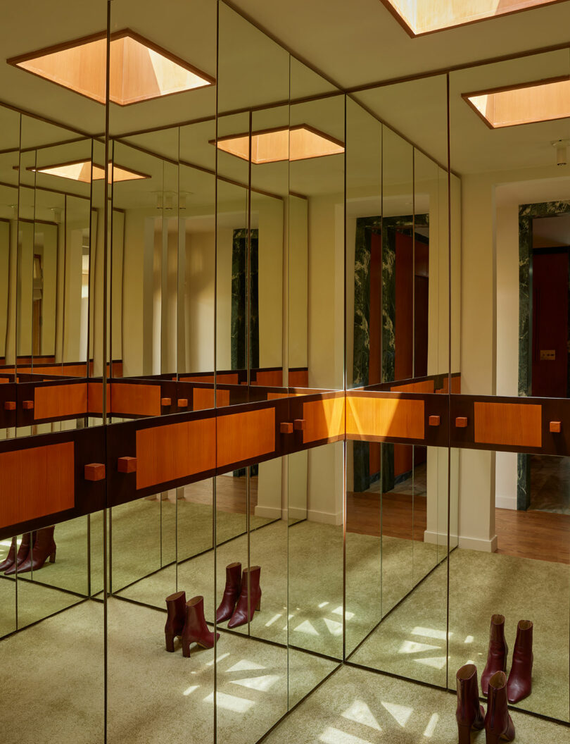 A room with mirrored walls and ceiling panels, beige carpet, wooden accents, and two pairs of burgundy boots placed on the floor.