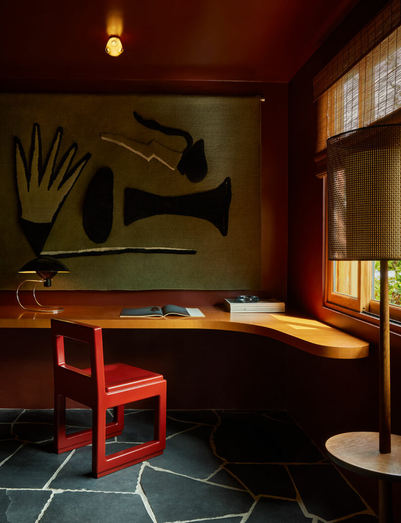 A red chair sits at a curved wooden desk under a large abstract wall hanging, with a lamp and an open book in a dimly lit room with dark tile flooring.