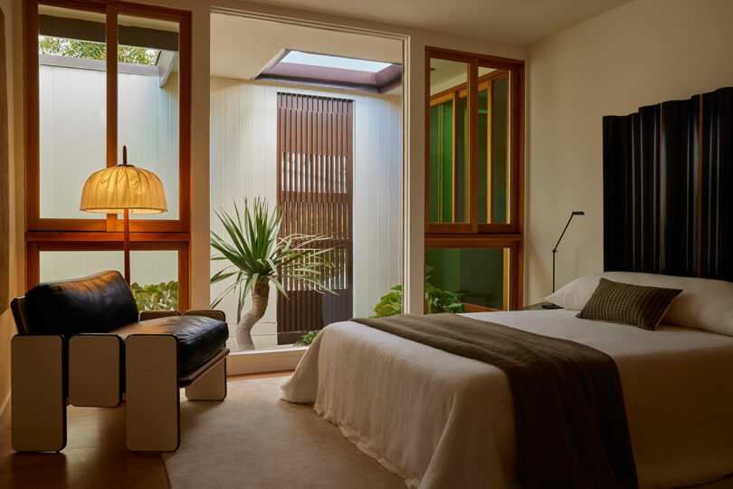 Modern bedroom with a bed, armchair, standing lamp, and large windows overlooking a small courtyard with plants and natural light.