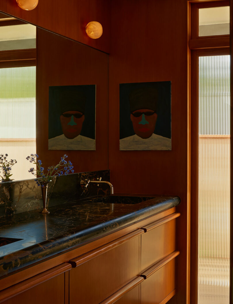 A bathroom with wood-paneled walls, a marble countertop with a sink, a small vase of flowers, and a painting reflected in a large mirror. Light enters through frosted glass panels.