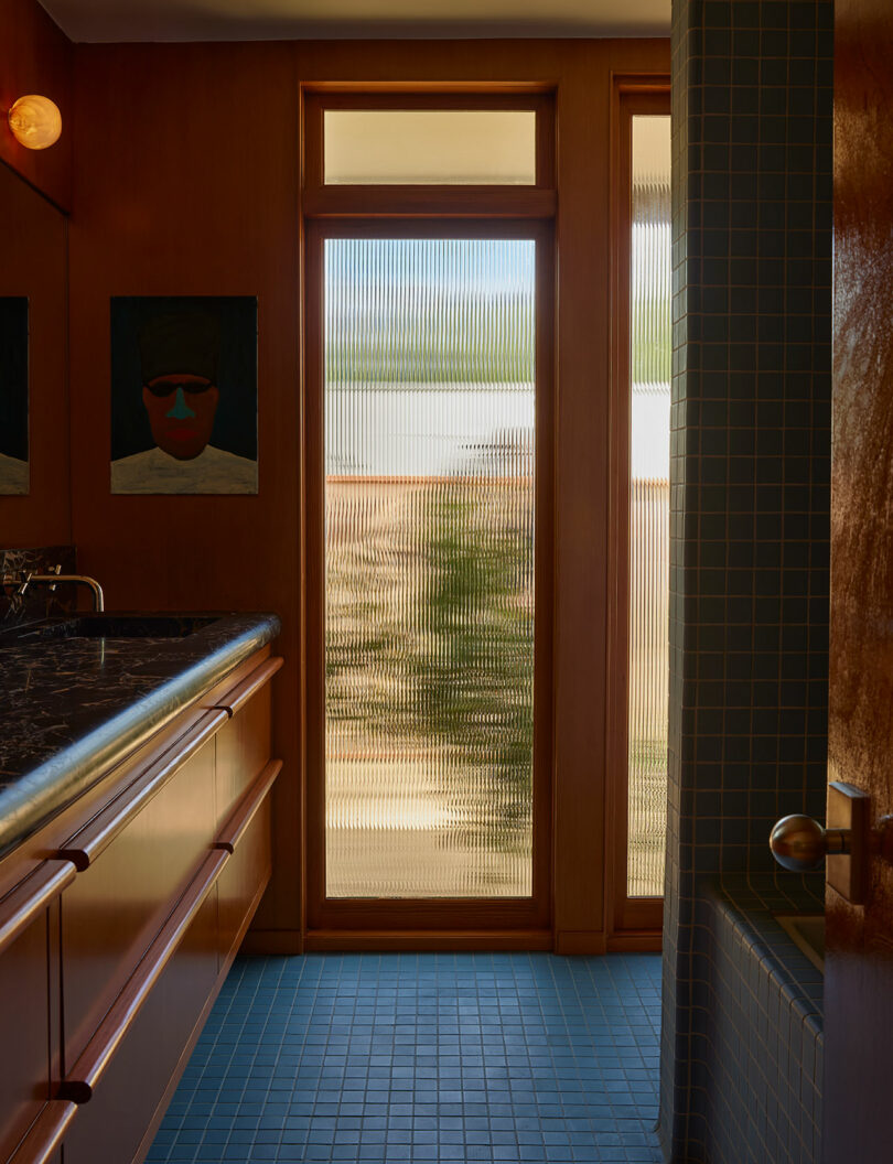A bathroom with wood-paneled walls, blue tile floor, a marble countertop, frosted glass windows, and a portrait hanging on the wall.
