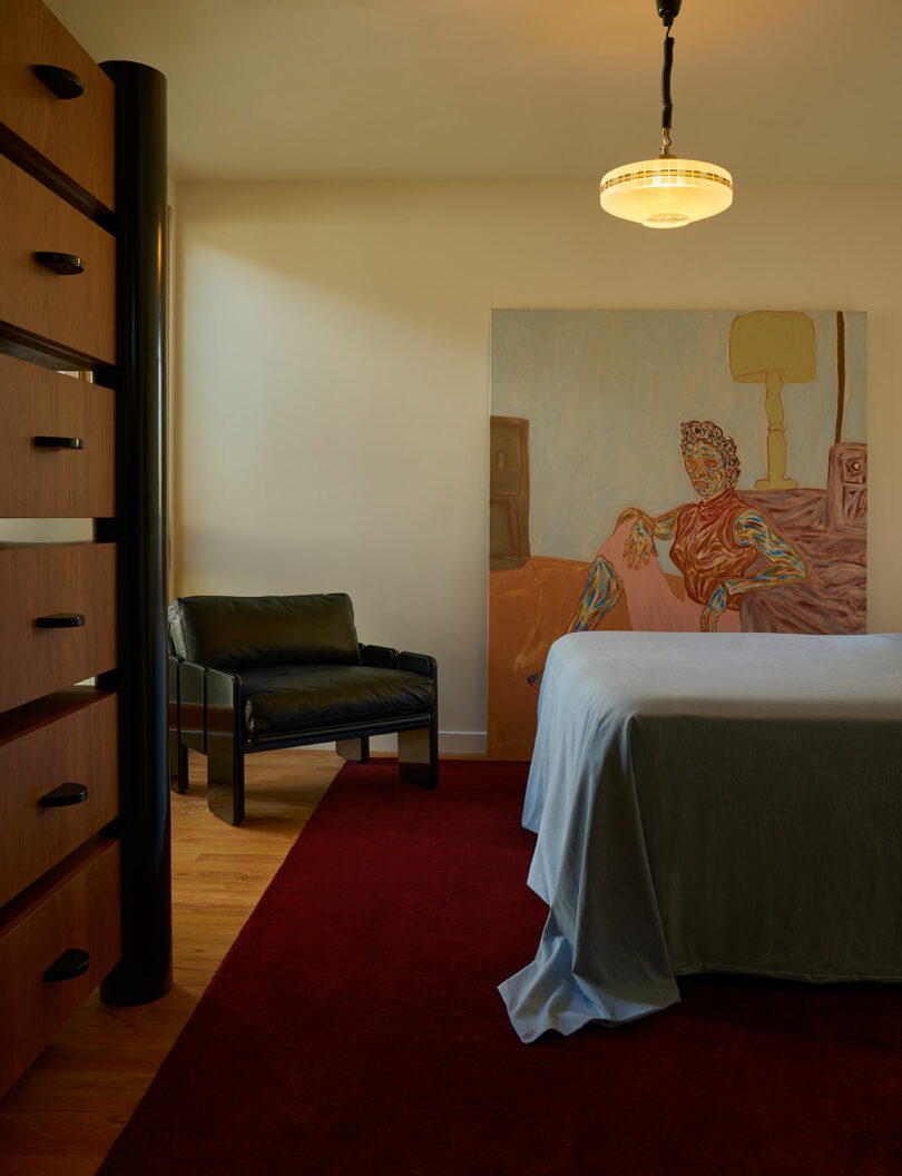 A minimalist bedroom with a black chair, a large dresser, a bed covered in a gray sheet, a red carpet, and a painting of a seated figure on the wall.