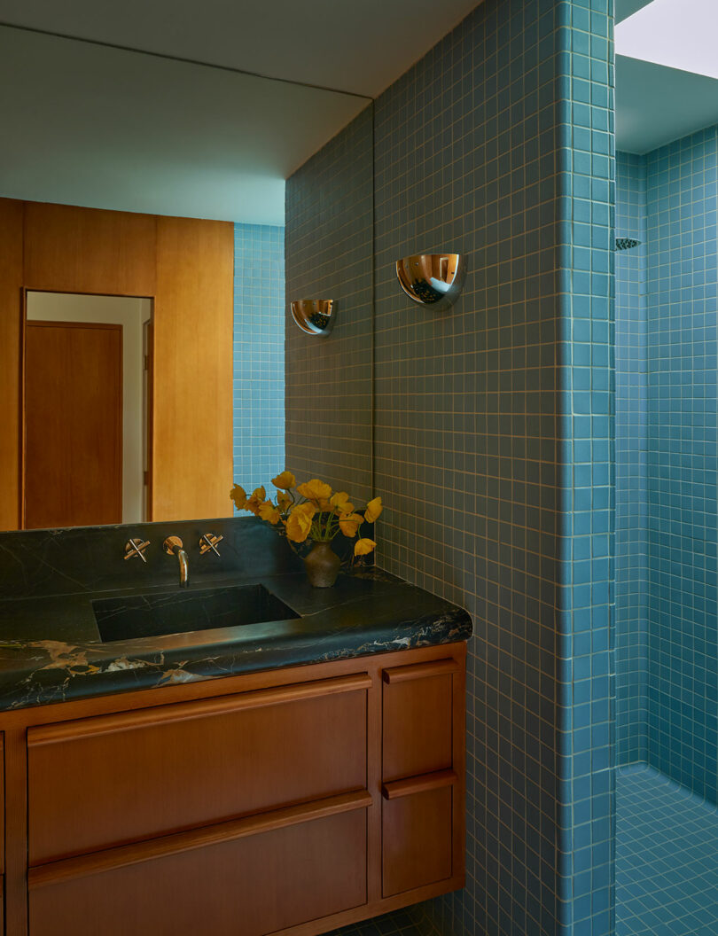 A modern bathroom with blue tile walls, a wooden vanity with a black marble countertop, a built-in sink, and a glass vase with yellow leaves.