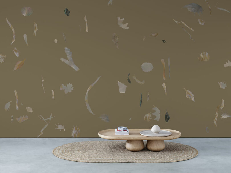 A low wooden coffee table with books and decor sits on a round woven rug in front of a brown wall with abstract shapes