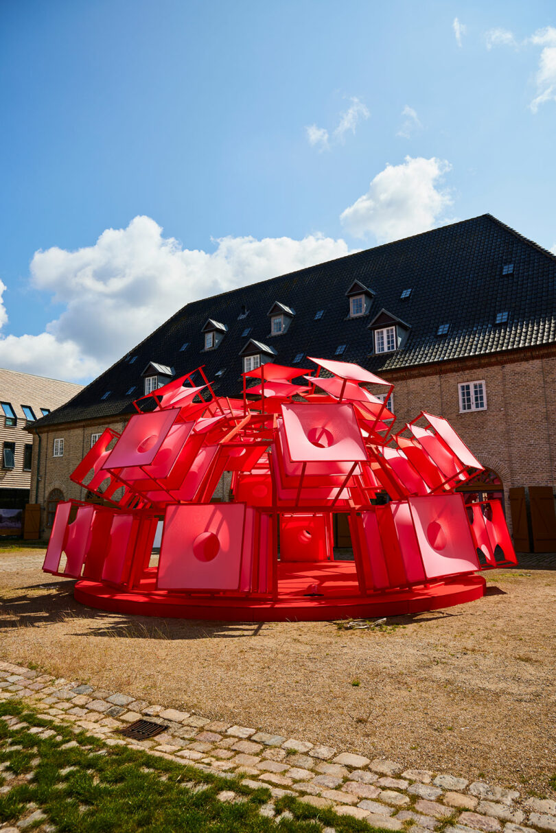 A large outdoor art installation made of red geometric panels arranged in a circular, dome-like structure stands in front of a brick building with a dark tiled roof.