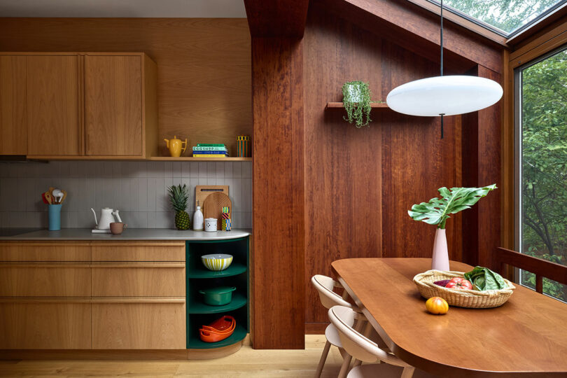 Cocina moderna con gabinetes de madera, estanterías abiertas, una mesa redonda de comedor con sillas, una claraboya y una canasta de frutas y verduras en la mesa.
