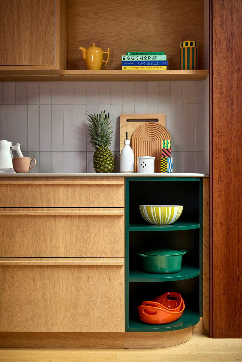 Una moderna esquina de cocina con gabinetes de madera, una piña, tablas de corte, tazones coloridos en los estantes y utensilios de cocina dispuestos en el mostrador y estanterías.
