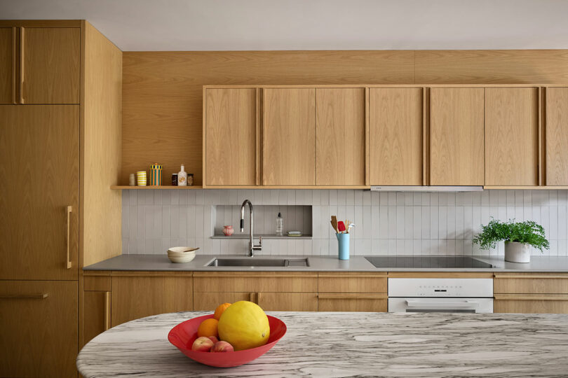 Cocina moderna con gabinetes de madera clara, encimera de mármol con un tazón rojo de frutas, fregadero de acero inoxidable, protector contra salpicaduras blancas y pequeña planta en maceta cerca de la estufa.