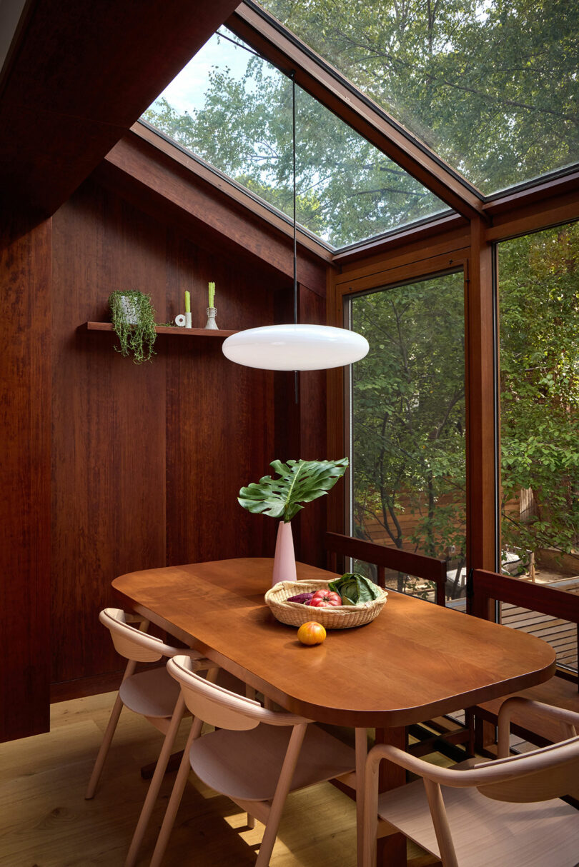 Una mesa de comedor de madera con cuatro sillas se encuentra en grandes ventanas y una claraboya. Una luz colgante blanca cuelga arriba, con un jarrón, un tazón de frutas y planta sobre la mesa. Los árboles son visibles afuera.
