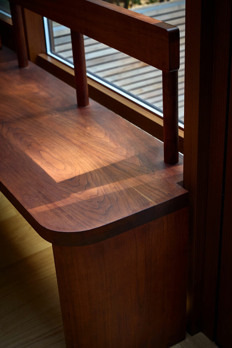 Un banco de madera con respaldo se coloca junto a una ventana, lanzando sombras en su superficie desde la luz natural.