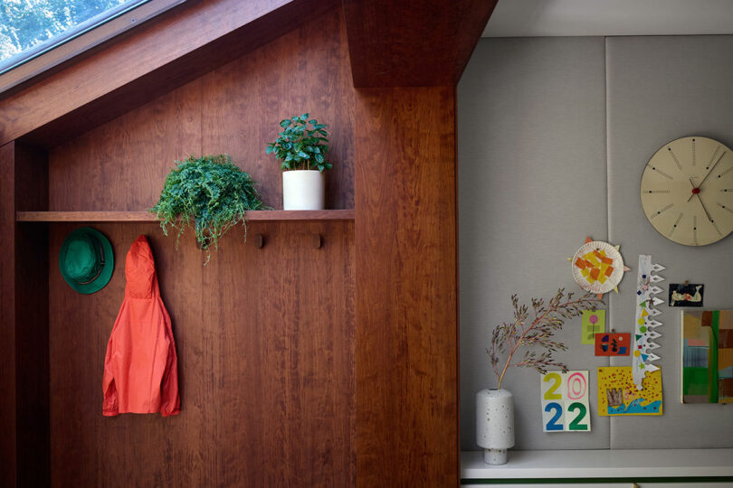 Una pared de madera con dos sombreros verdes, una chaqueta naranja y dos plantas en macetas; La pared adyacente muestra un reloj, jarrón con ramas y coloridas obras de arte.