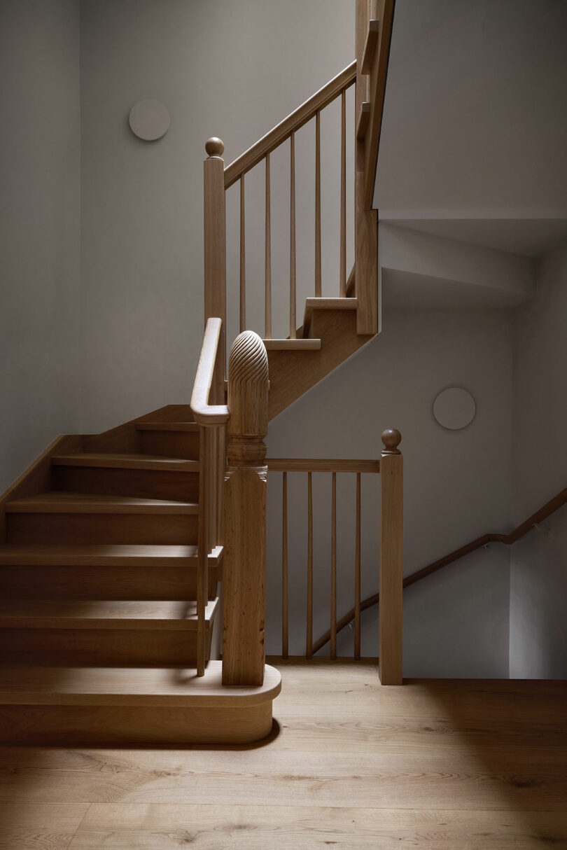 Escalera de madera con barandillas simples y postes redondeados, colocados contra paredes blancas lisas en un espacio interior bien iluminado.