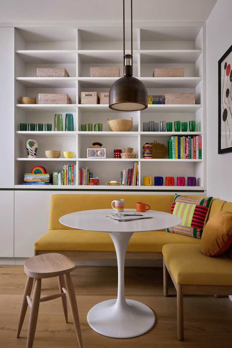 Un rincón de comedor moderno con un banco tapizado amarillo, mesa redonda blanca, taburetes de madera, tazas coloridas, libros y artículos decorativos en estantes abiertos contra una pared blanca.