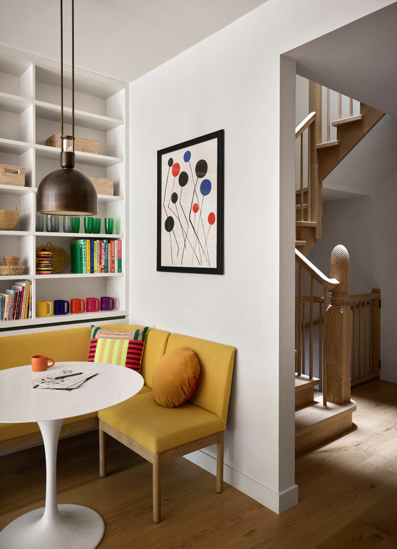 Un rincón de comedor moderno con un banco amarillo, mesa blanca redonda, tazas coloridas, estantes incorporados, obras de arte abstractas y una escalera de madera en el fondo.