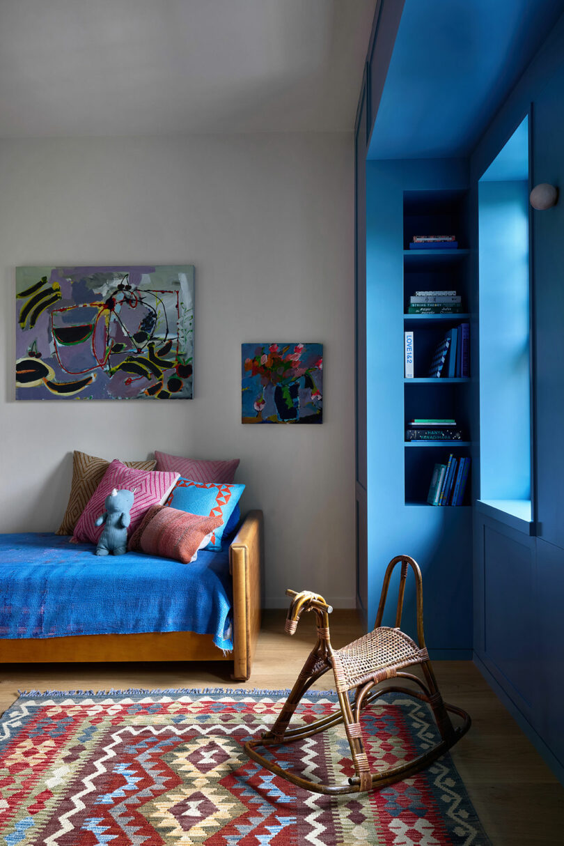 Una habitación para niños colorida con una cama azul, una alfombra estampada, un caballo mecedora de mimbre, estantes empotrados azules y pinturas abstractas en la pared.