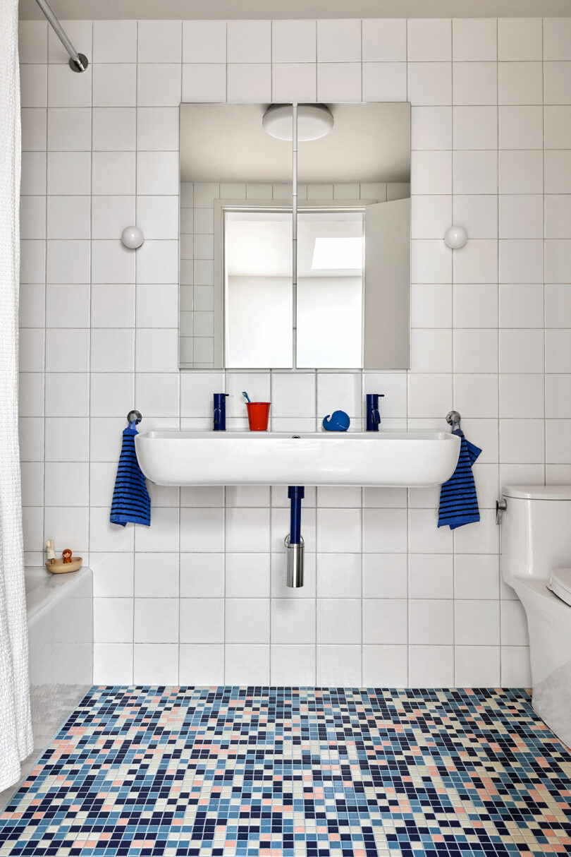 Baño moderno con paredes de azulejos blancos, un fregadero rectangular, dos espejos, azulejos de mosaico azul y blanco, toallas azules rayadas y accesorios mínimos.