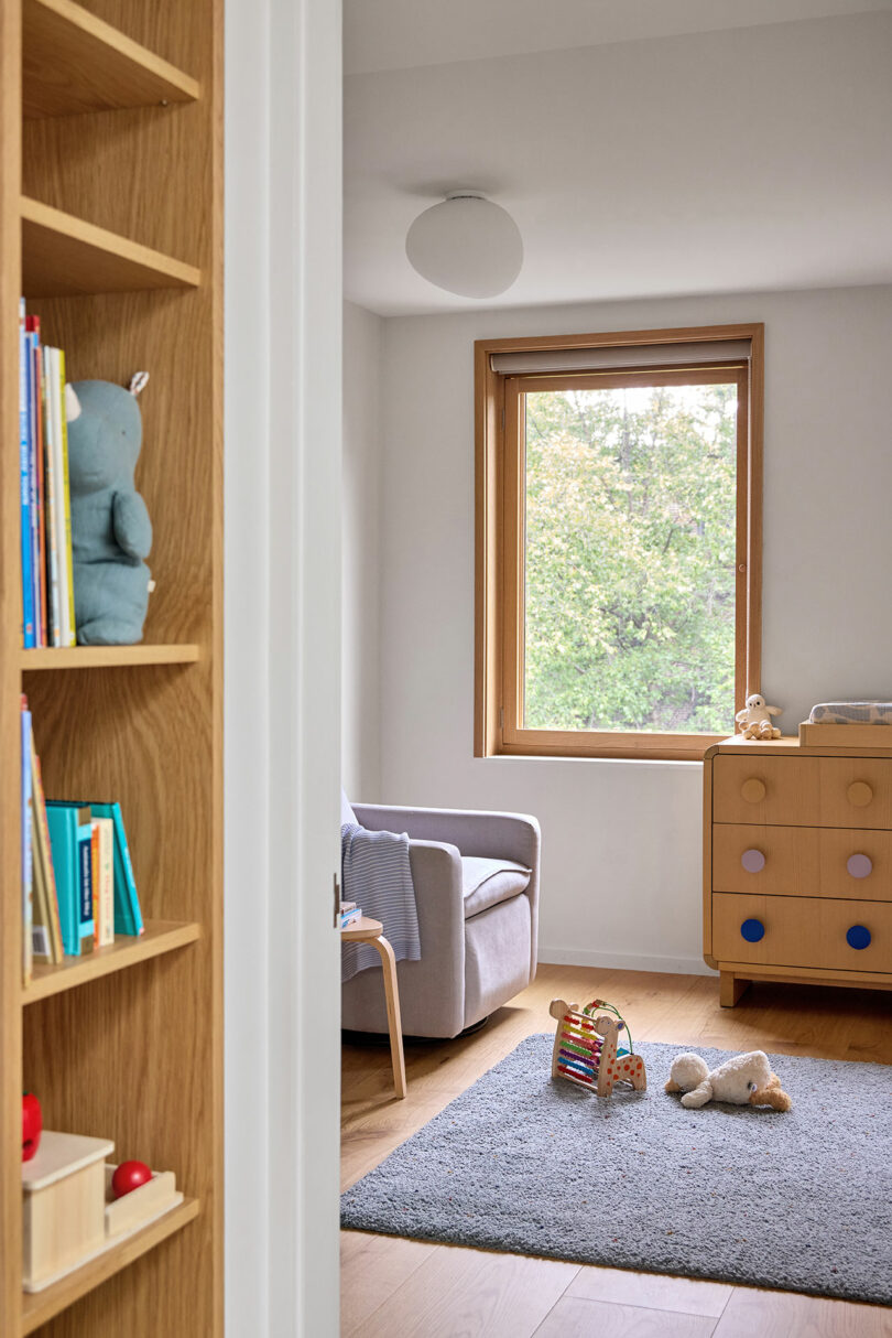 Una habitación infantil brillante y ordenada con un sillón gris, tocador de madera, ventana, juguetes en una alfombra gris y una estantería parcialmente visible en primer plano.