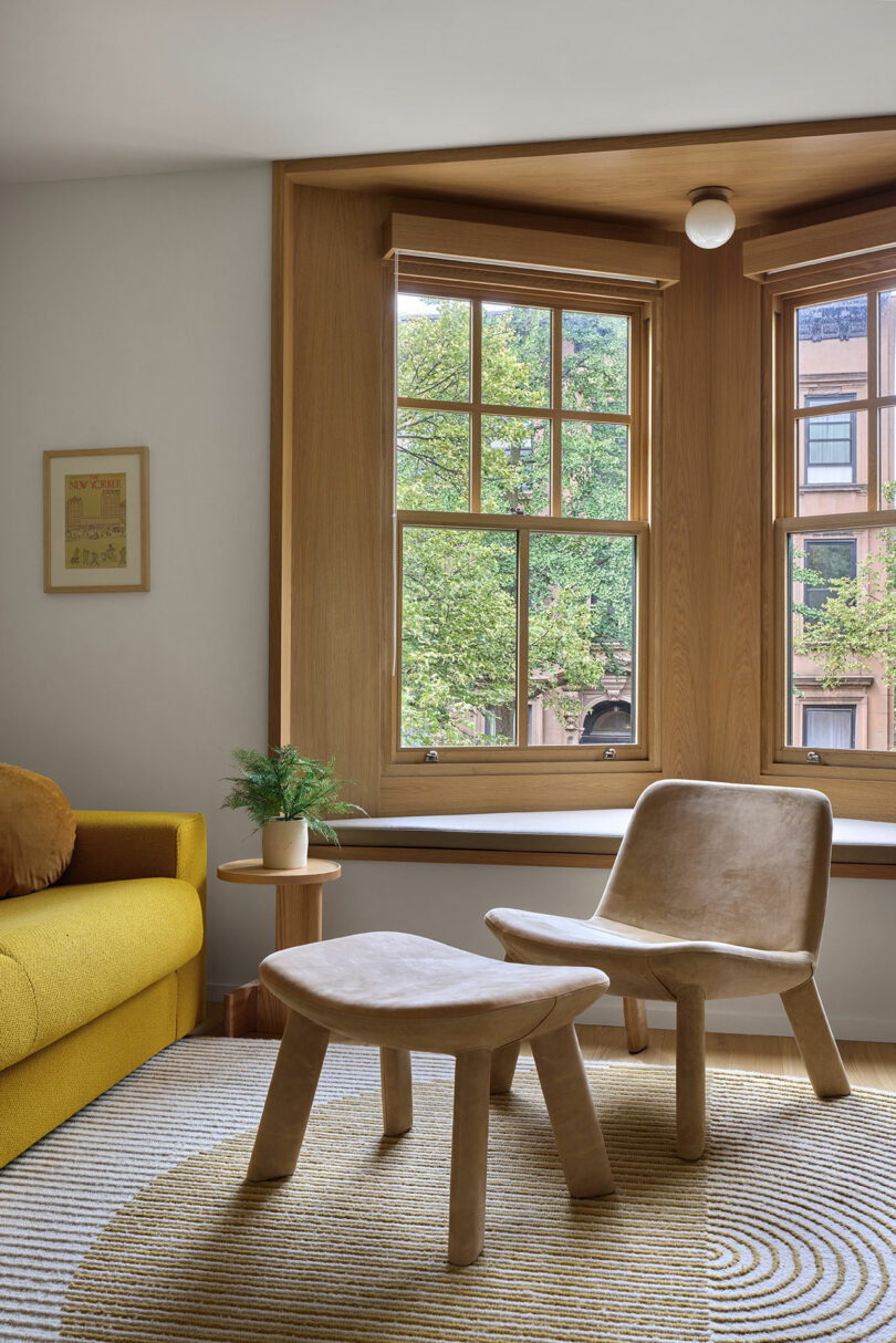 Un acogedor rincón de lectura con sillas de madera, una pequeña mesa auxiliar y un asiento de ventana da a los árboles frondosos a través de grandes ventanas de laurel. Un sofá amarillo es visible a la izquierda.