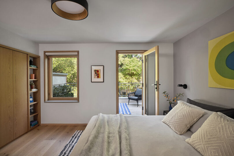 Un dormitorio moderno con una cama cuidadosamente hecha, pisos de madera, estantes incorporados y una puerta abierta que conduce a una terraza con asientos al aire libre.