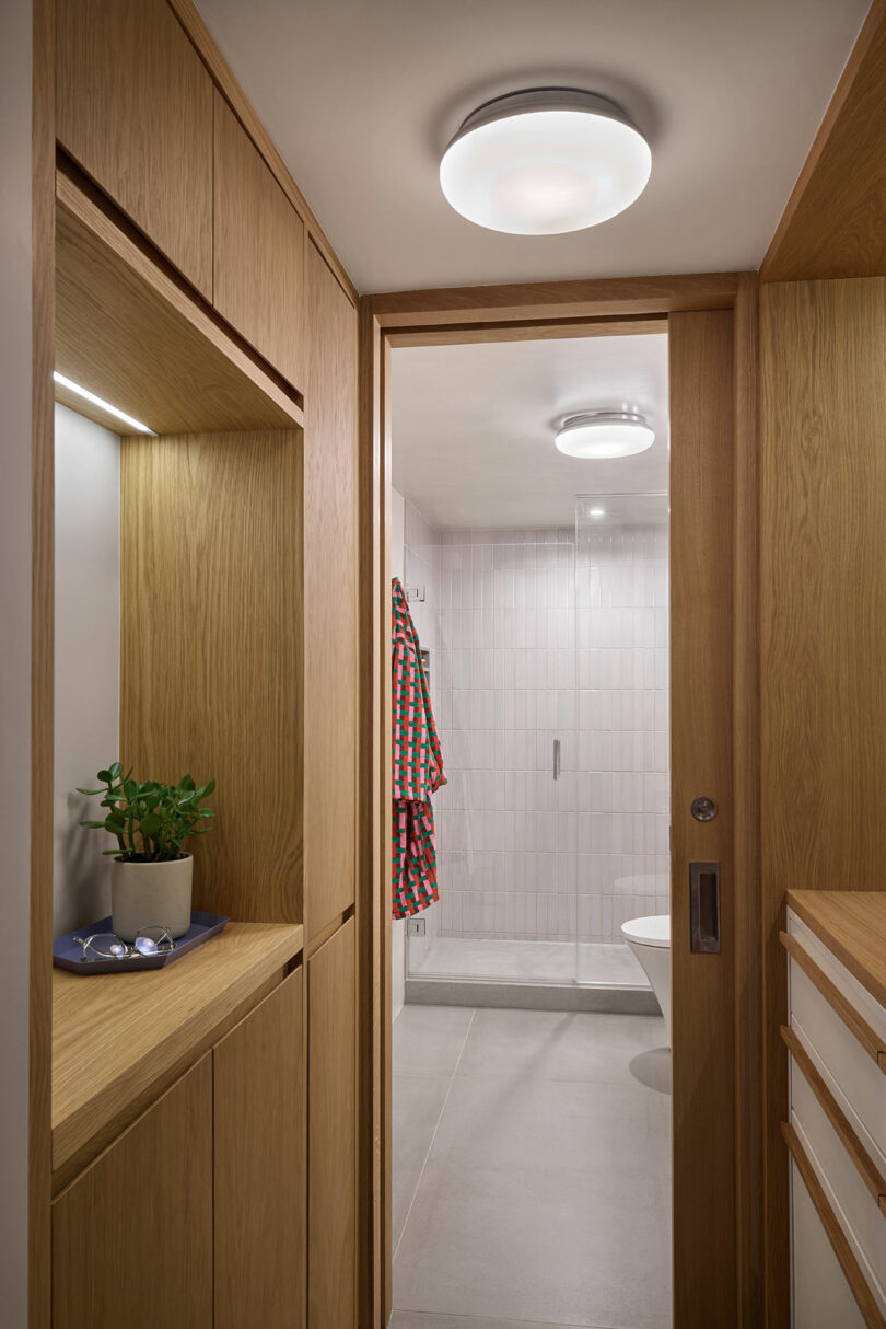 Un pasillo con gabinetes de madera incorporados conduce a un baño con ducha de vidrio, paredes de azulejos y una bata a cuadros colgando en la pared.