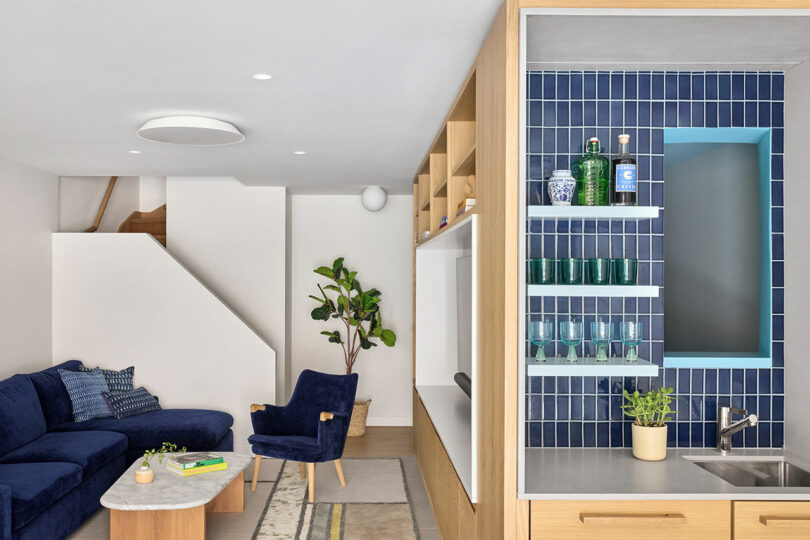 Sala de estar moderna con un sofá seccional azul marino, silla de madera, mesa de café de mármol y un área de cocina con baldosas azules, estantes abiertos y un pequeño fregadero.