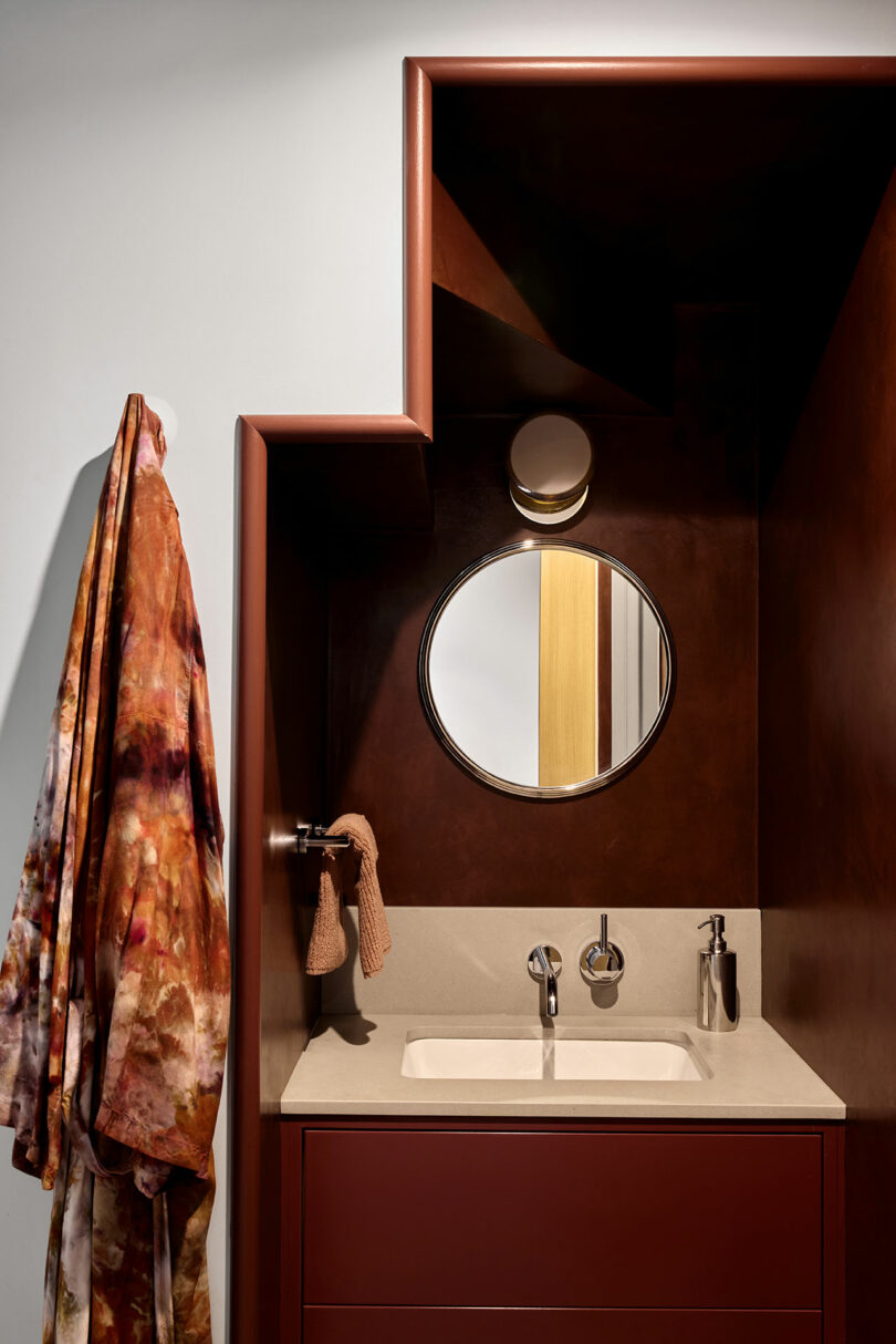 Un fregadero de baño moderno con un espejo circular, grifo montado en la pared, toalla pequeña, dispensador de jabón y una túnica colorida colgando de un gancho.