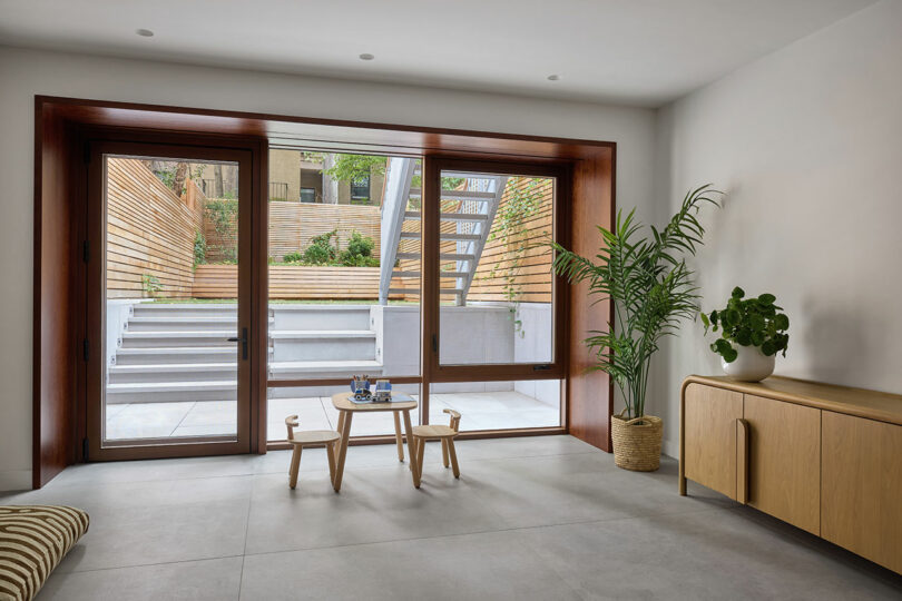Una habitación moderna con grandes puertas de vidrio que se abren a un patio trasero en terrazas, con una pequeña mesa de madera con taburetes, plantas y un aparador de madera ligera.