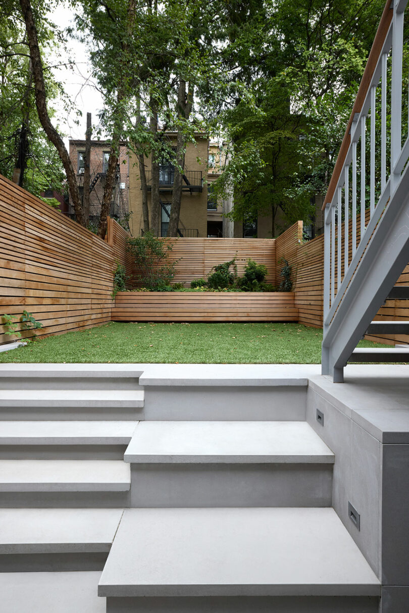 El patio trasero moderno con pasos de concreto blanco que conducen a un área cubierta de hierba, cercas de privacidad de madera, macetas con arbustos y árboles circundantes.