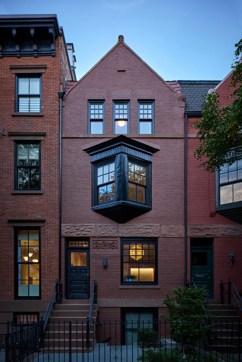 Una casa de ladrillo con una ventana salada, puerta negra, escalones delanteros y una cerca de hierro, flanqueada por edificios vecinos similares al anochecer.