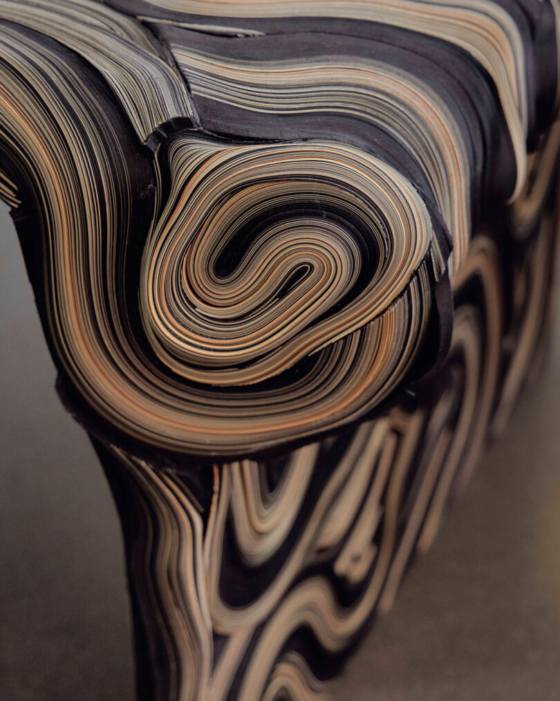Close-up of a sculpted object with layered, swirling patterns in black, brown, and beige, reminiscent of stratified wood or paper—characteristic of the Fragmenti series by Daniele Papuli.