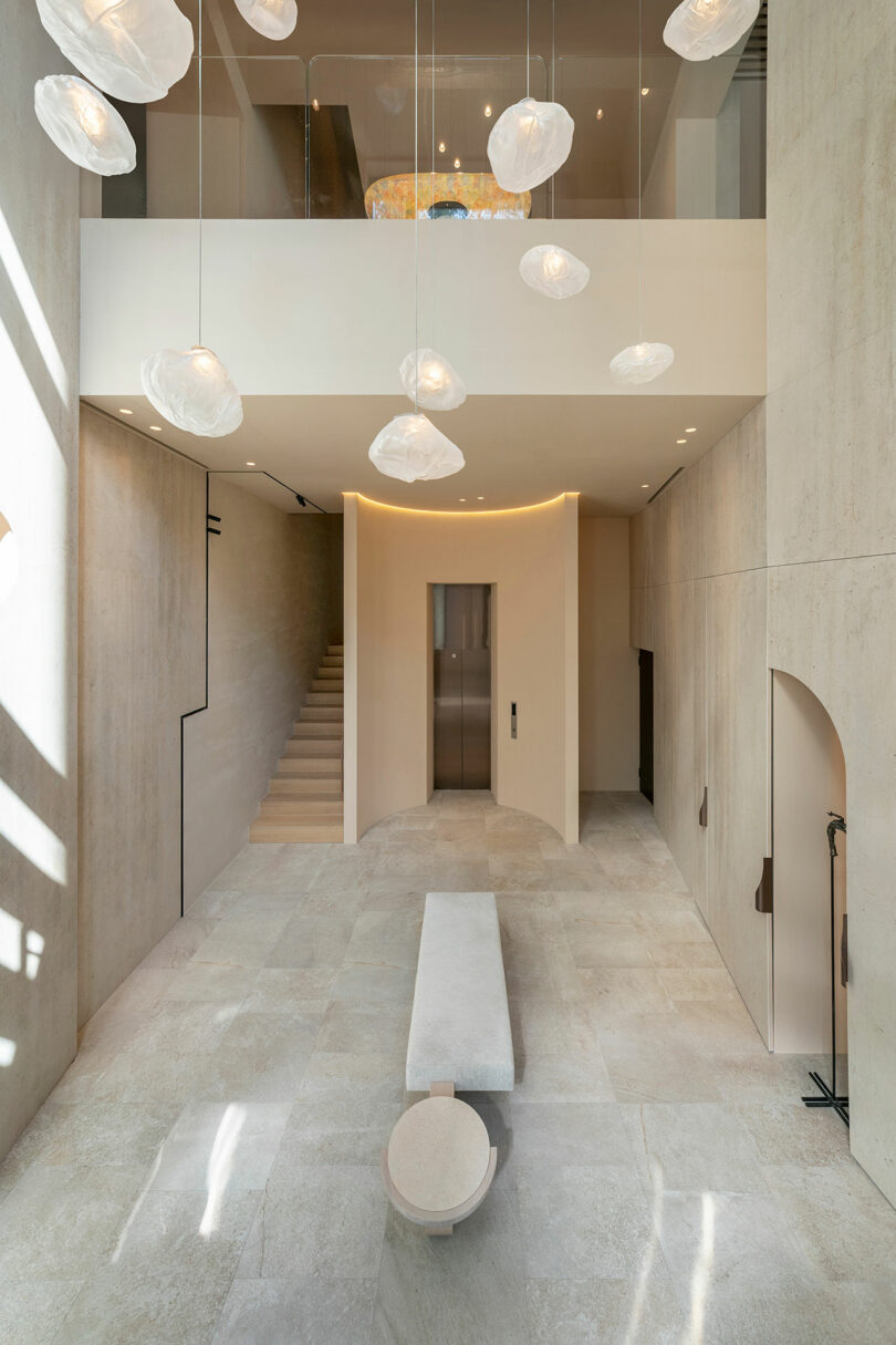 Minimalist interior with stone floors, a bench and round table, floating stairway, central elevator, and organic-shaped pendant lights hanging from a double-height ceiling.