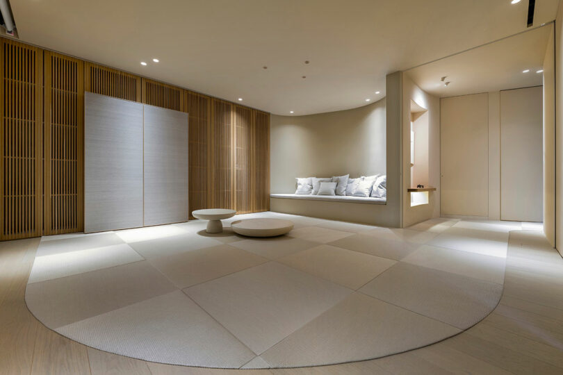 Minimalist room with light wood walls, low round tables, large tatami mats, built-in seating area with cushions, and soft ambient lighting.