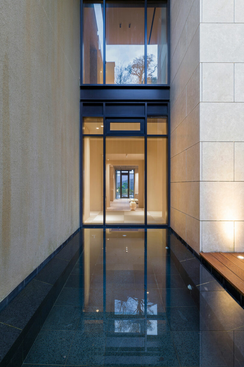 A modern building entryway with large glass doors, beige stone walls, and a reflective water feature leading to an illuminated interior space.