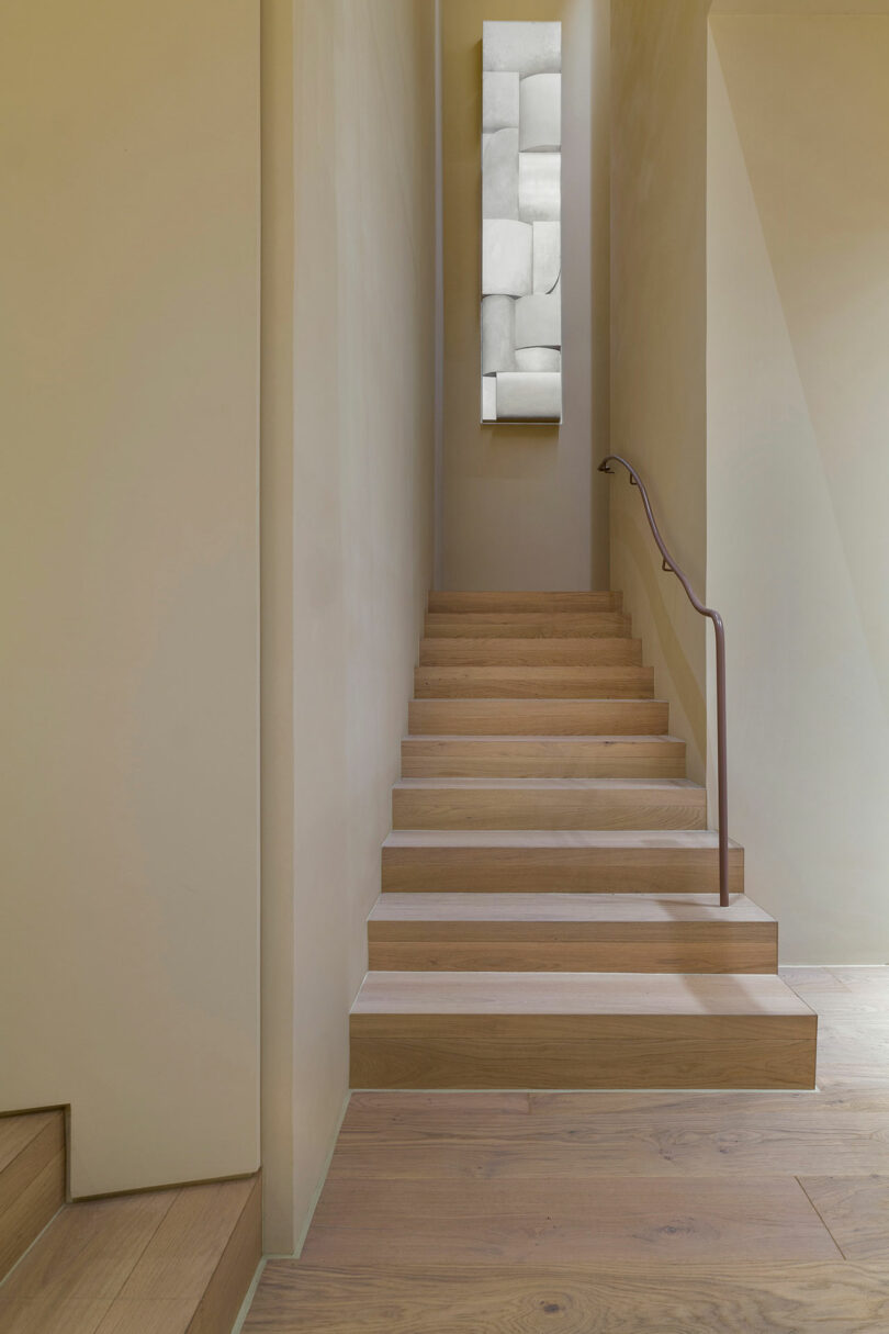A minimalist wooden staircase with a simple handrail leads up to a window displaying geometric frosted glass panels.