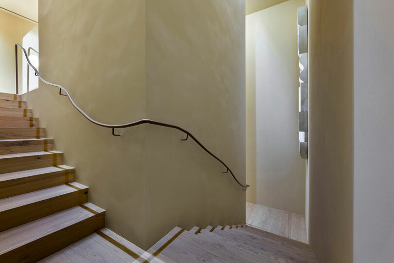 Wooden staircase with a simple, curved metal handrail on a beige wall, leading up and turning around a corner in a minimalist interior space.
