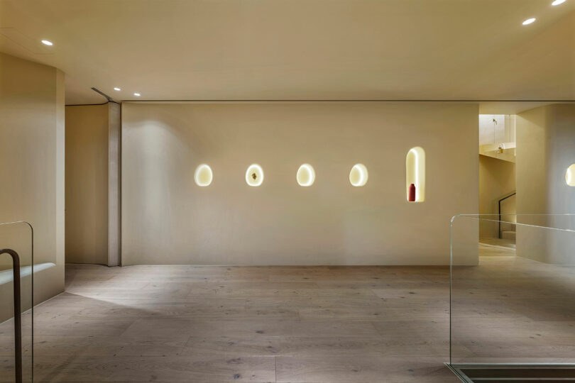 Minimalist interior with a light wood floor, beige walls, and five oval niches illuminated on the wall, each containing a small object. Stairs are visible in the background.