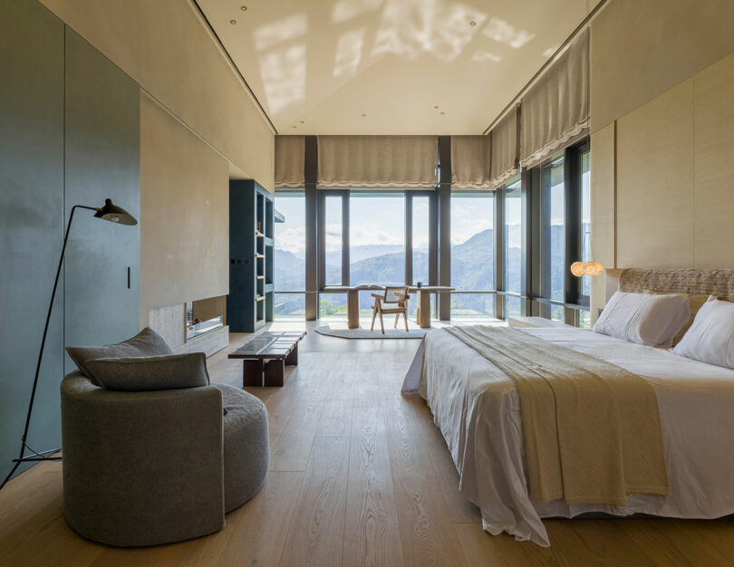 Modern bedroom with large windows, a double bed, grey armchair, and a scenic mountain view outside. Light wood flooring and neutral décor create a bright, spacious atmosphere.