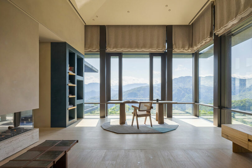Modern minimalist room with large windows, a wooden chair and table, built-in shelves, and expansive mountain views in the background.