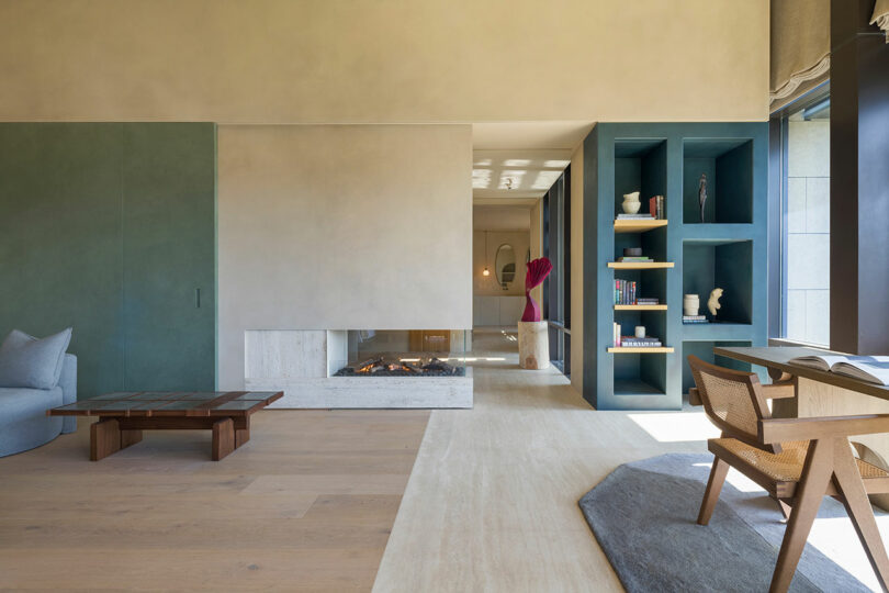 Modern living room with a minimalist fireplace, built-in shelving, a wooden coffee table, armchair, desk, and large window letting in natural light.